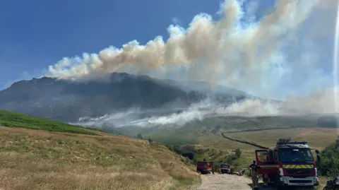 Smoke billows above hills and moorland with a fire engine parked at the bottom