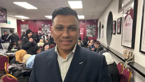 Kate Bradbrook A man in a navy blazer and white striped shirt stands in a busy room.