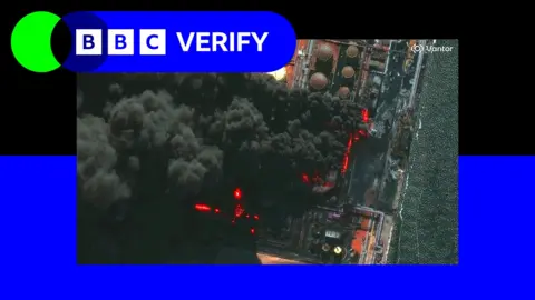 A large smoke plume rises from oil tanks, there are visible flames in the satellite image and the sea nearby is visible
