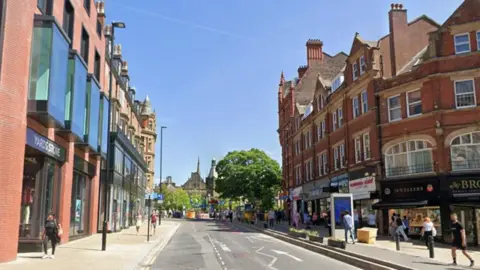 A road with shops in four-storey Victorian era buildings on either side. No vehicles are on the road. People walking on both sides of the road. The sky is blue.