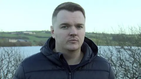 A man is looking into the camera. A lake and fields are behind him. He's wearing a navy coat. 