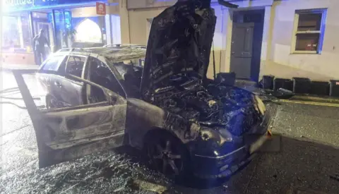 Brixham Fire Station A car - possibly a Skoda - with its bonnet and a car door open and the charred remains of the engine and the interior of the car along with flame marks on the exterior suggesting that the car has been on fire, with a house behind along with a line of what appear to be household waste vestibules which appear to of the food caddy genre.