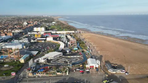 A wide aerial view of Skegness showing a large sandy beach stretching along the coastline beside calm blue-grey water. Next to the beach is a busy seafront area with amusement rides, go‑kart tracks, and various buildings, including arcades and attractions. Further inland, rows of houses and streets extend into the distance. The scene is bright and clear, with the coastline curving away towards the horizon.
