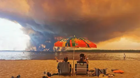 Maximilien Lamy/AFP via Getty Images Beachgoers rest on the beach in Lacanau as clouds of smoke rise into the sky from a wildfire in the distance.