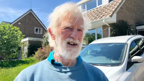 A man stood in front of a house with a white car parked on a drive. He has white hair and beard and is wearing a blue sweatshirt with a light blue polo top underneath. He has a gap in his top front teeth. There is also a neighbouring house behind him.