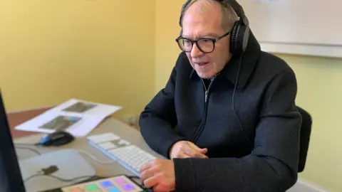 A man wearing a dark jacket, headphones over his ears is sitting at a desk with a computer and mixing desk in front of him