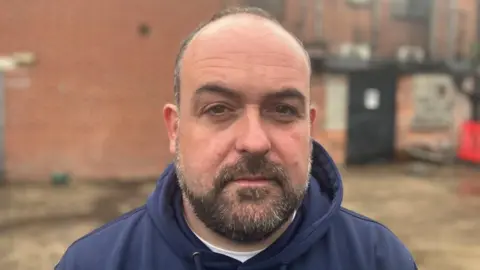 McCusker is looking directly at the camera. He has brown/grey facial hair and thin grey hair. He is wearing a navy hoodie.