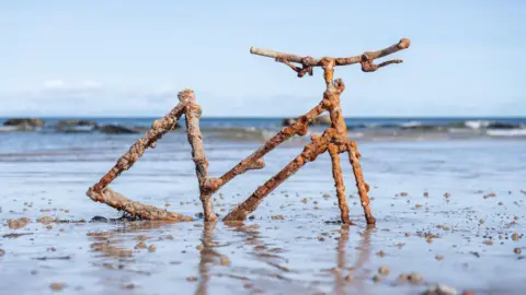 Ian Knight A rust-covered metal sculpture shaped like a bicycle stands partially embedded in wet sand on a beach, with calm sea water and a clear blue sky in the background.