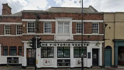 A two storey building with a white facade on the ground floor and brick on the first. The ground floor has a sign above the windows and doors that says The Victoria Vaults in black