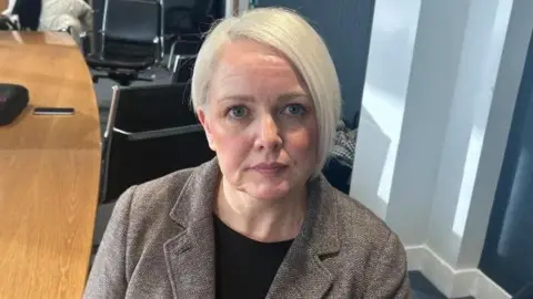 Detective Inspector Noelle Gray has short light blond hair, in a bob hairstyle. She is looking directly at the camera with a straight face expression. She is wearing a light brown blazer and a clack top underneath. 