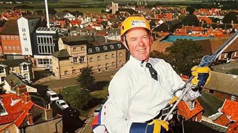 John Huggins, wearing a hard helmet, a white shirt and tie with sturdy safety gloves, leans back against the ropes as he starts an abseil down the lighthouse. Mr Huggins is very high up above the rooftops of houses and commercial buildings with the water tower and fields in the background.