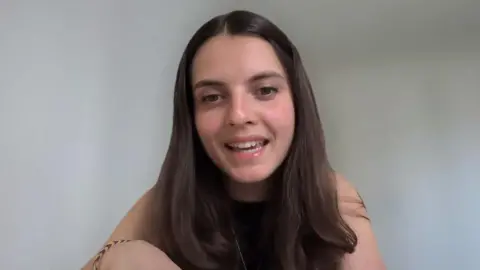 A lady with brown hair and a black top talks on a Zoom meeting.