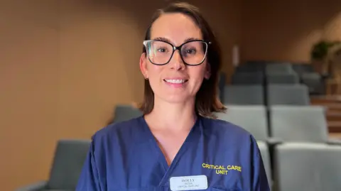 Holly is wearing blue clinical scrubs stands in front of rows of blue auditorium-style seats. The scrub top features the embroidered text “CRITICAL CARE UNIT” and a staff name badge. The setting appears to be a lecture theatre or hospital education space with softly blurred seating in the background.