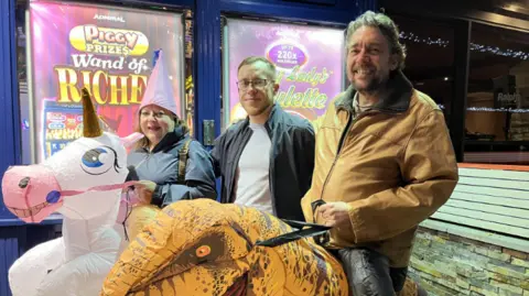 A woman and two men stand in a row wearing fancy dress. The woman is wearing a blow up pink unicorn, along with a large pointy pink hat. The man in the middle is wearing a white top and a jacket. The second man is wearing a blow up dinosaur costumer. They are all smiling at the camera.