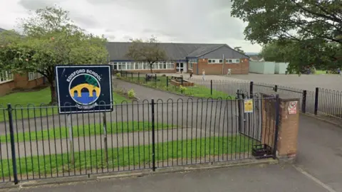 Google Streetview image of Edisford Primary School in Clitheroe. It is a single-storey building with a large playground and a large tree to the right and railings in front. To the left is another tree behind a sign that says "Edisford School".