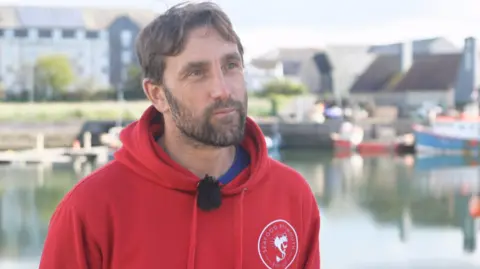A man wearing a red hoodie. He is stood in front of a quay with boats in the background.