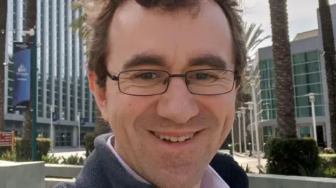 RNIB John Paton has short brown hair and is wearing black-rimmed glasses. He is wearing a pink shirt and blue fleece. John is smiling at the camera. He is standing in a city with a number of palm trees behind him. 