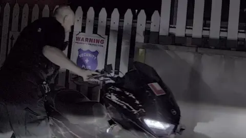 A bald police officer dressed in black uniform holds onto the handlebars of a black damaged moped while looking down at it. The headlight is on as it is dark. Behind the moped is a white fence with a warning sign on it.