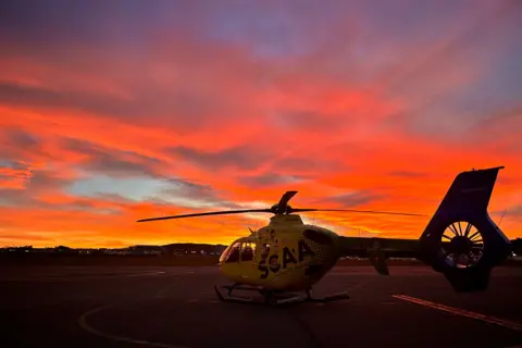 Suzanne Watson A yellow helicopter sits on the tarmac with a sunset behind it. The sky is orange, red and yellow.