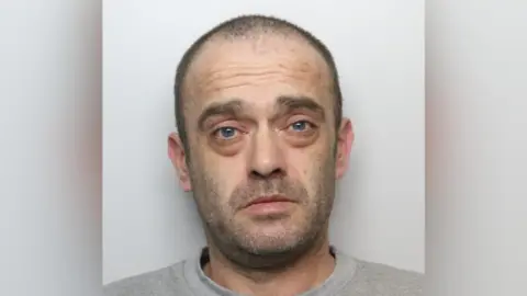 A police mugshot of Ashley Asquith. He has stubble and short receding hair. He is wearing a grey jumper.