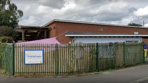 A nursery building in Gloucester built out of red brick with a grey rendered entryway. The building sits behind green fencing - which connects to the building with a pink play canopy - and has a sign that reads "Dingley's Promise"