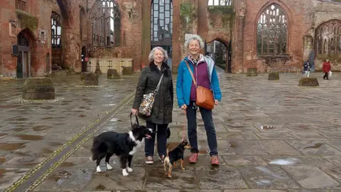 Clare Slator Two women stand with two dogs in front of the red sandstone ruins of a Gothic-style cathedral, featuring tall arched windows and spires. The cathedral is roofless, with only its outer walls intact. The ground is paved with large stone slabs. Other visitors are visible in the background, exploring the site.