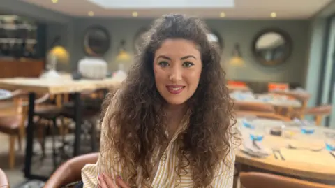 Alexandra Rotaru looking at the camera sat in the restaurant of the Blonde Hedgehog. Alexandra has brown curly hair, and is wearing a white and yellow stripey shirt.
