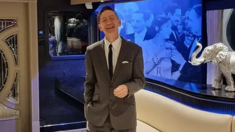 Facebook Thomas Flight dressed in a grey suit and black tie. He is standing on his own in front of a display showing an old photograph and a silver elephant statue.