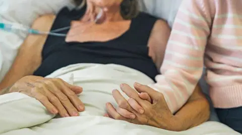 In the bed of a hospital, a getty image patient is holding his hand with a loved one who is visiting.