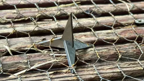 Metal throwing star resting on chicken wire laid over stacked, weathered wooden logs