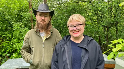 Lee and Emma stand outside at the collective. They both wear zip-up coats and Lee wears a back hat.