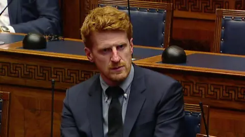 Matthew O'Toole has red hair parted to the side and a thin red beard. He is sitting in the assembly chamber