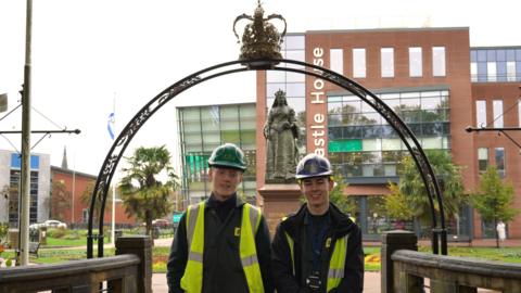 Two apprentices from IAE stand in front of the decorative archway at the entrance to Queen's Gardens. 