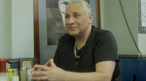 Hussein is seen wearing a black T-shirt sits at a table inside a café, with condiment bottles and menus on the table in front of him.