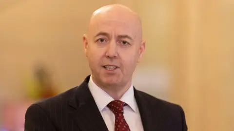 PA Media Jon Burrows is photographed looking toward the camera while speaking. He is bald and wearing a dark pinstriped suit, white shirt and red tie.