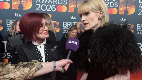 A woman with red hair and a woman with blonde hair and a fur coat, stand on a red carpet speaking into a microphone.