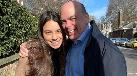 PA Mike and Hannah Lynch smile for the camera on the street in front of some hedges and a low brick wall. Mike put his arm around Hana's shoulder.