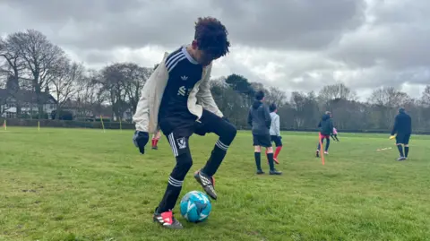 Claire Hamilton/BBC A young boy, with brown curly hair and wearing a cream zip-up training jacket over a black replica Liverpool FC football kit, dribbles a blue Mitre football on a grassy field. Other boys can be seen training behind him. 