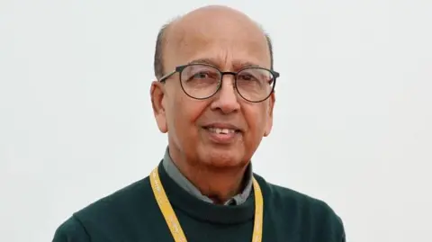 Prof Ahmed has heavily receding dark hair and is wearing black round spectacles on his face, a dark-green jumper over a sage-coloured shirt.