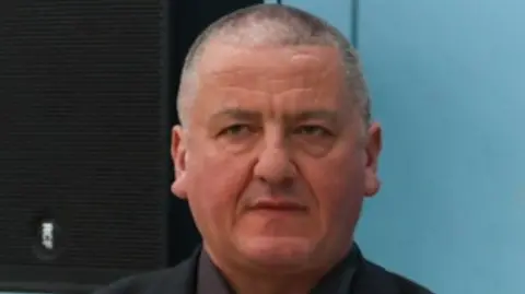 Andrew Harrison is wearing a black suit with a dark red tie with a light blue Reform rosette. He has close cropped grey hair.