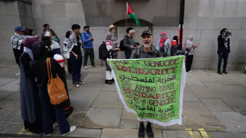 Pa Media Group Protests outside old Bailey, London. Many are wearing a Palestine White fan and misleading the Palestine Flag. A man stands in front of catching green signs and words 'stops, solidarity, stops the hand of Israel, solid, stops the ginuditity'.