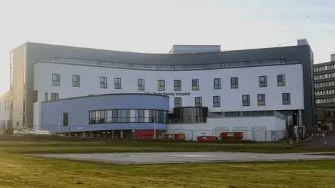 BBC The hospital is a large, four storey white and grey building, part of the building is blue and extends from the front of the hospital. 