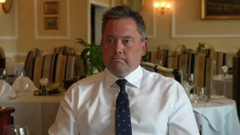 Peter Brend, director of Brend Hotels, sits in the dining room of a hotel. He is wearing a white shirt with a navy blue tie which has silver patterns on it. He has short greying hair.