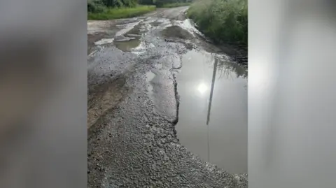 A damaged road covered with large potholes filled with water and grass verges on either side.