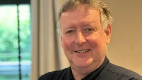 A man wearing a black shirt is looking at the camera and smiling. He has grey hair and is standing indoors.