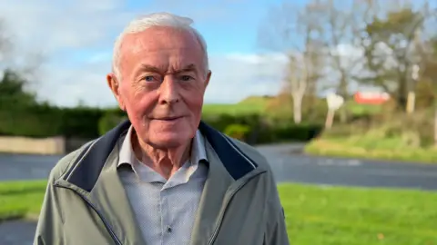 An older man with white hair looks directly into the camera, standing in front of a rural road and grass verge, with trees and fields in the background. 