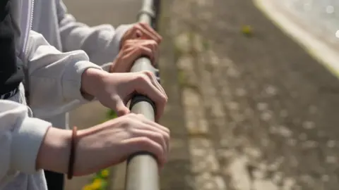 Two people wearing grey tops hold on to a metal fence pole near a lake