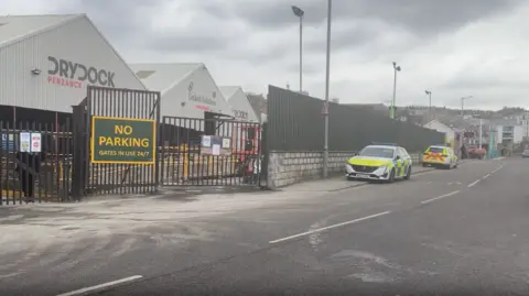 Penzance Dry Dock has three grey buildings next to a main road. There are two police cars parked on the pavement. A sign on the gates reads NO PARKING GATES IN USE 24/7. 