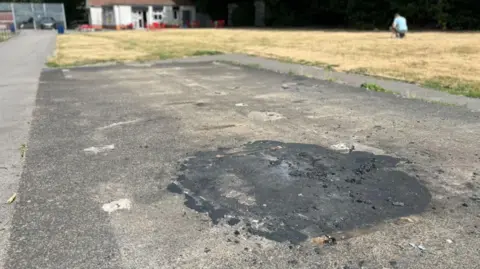 LDRS A patch of concrete with a large darker patch on it. In the background a dry, yellowing patch of grass and a white building can be seen.