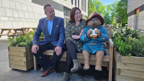 Paddington Bear statue dressed in his trademark red hat and blue coat while eating a marmalade sandwich. He is sitting on a bench with two wooden planters filled with flowers beside him. A man dressed in a blue suit and a woman in a green dress sit beside Paddington